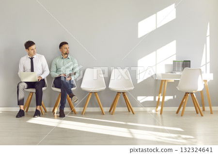 Two men sitting by office wall, waiting for job interview and looking to side at table with laptop Two men sitting by office wall, waiting for job interview and looking to side at table with laptop 132324641