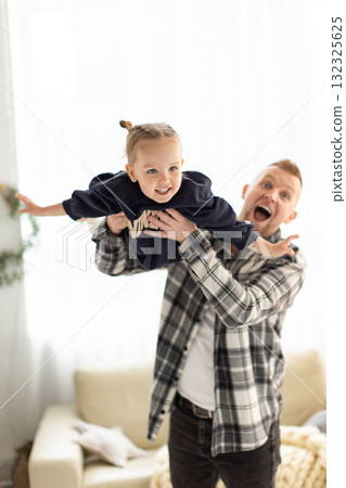 A happy father holds his young daughter up in the air, both smiling and enjoying a playful moment indoors. A happy father holds his young daughter up in the air, both smiling and enjoying a playful moment indoors. 132325625