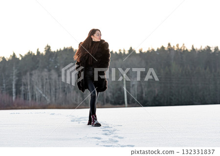 a cheerful girl in a warm fur coat walks across the open field leaving footpath in the snow 132331837