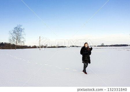 a cheerful girl in a warm fur coat walks across the open field leaving footpath in the snow 132331840