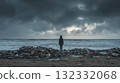 Solitary Figure Stands on Polluted Beach Under a Stormy Sky Facing the Rough Sea 132332068