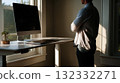 Man Stands By Window in Home Office With Computer, Laptop, and Plant on Desk 132332271