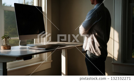 Man Stands By Window in Home Office With Computer, Laptop, and Plant on Desk Man Stands By Window in Home Office With Computer, Laptop, and Plant on Desk 132332271