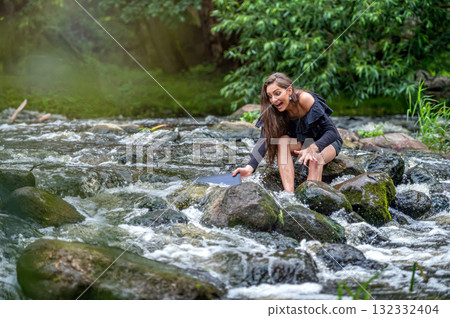 female freelancer sitting on a rock in the river and dropping her laptop into the water 132332404