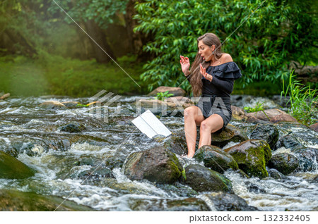 female freelancer sitting on a rock in the river and dropping her laptop into the water 132332405