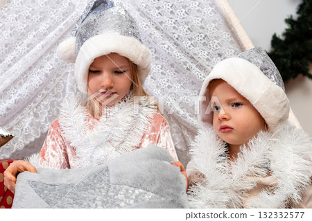 young children in silver Christmas hats sit between gift boxes and toys in a decorated house young children in silver Christmas hats sit between gift boxes and toys in a decorated house 132332577