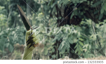Witch hand with a long black nail holding a knife with a haunted forest background. Zombie, devil, evil, satan, or monster hand. Concept of Spooky Halloween 132333935