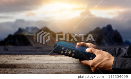 Prayer holding the blue book of the holy bible on a wooden table with a landscape view and a dark dramatic sky background. Concept of Christianity, religious, faith, Jesus, or belief 132334099