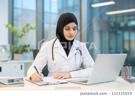 Muslim female doctor wearing a hijab and stethoscope, carefully writing notes and using a laptop while sitting at her desk in a contemporary healthcare office environment Muslim female doctor wearing a hijab and stethoscope, carefully writing notes and using a laptop while sitting at her desk in a contemporary healthcare office environment 132335059