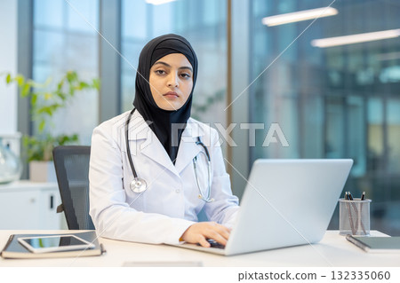 Muslim female doctor wearing a hijab and white lab coat, focusing on her laptop while sitting at a desk in a modern medical office setting, representing healthcare and technology 132335060