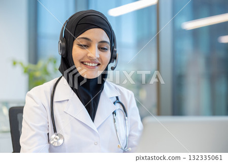 Friendly muslim doctor wearing headset and hijab working on her laptop, providing telehealth services and smiling at a patient on a video call Friendly muslim doctor wearing headset and hijab working on her laptop, providing telehealth services and smiling at a patient on a video call 132335061
