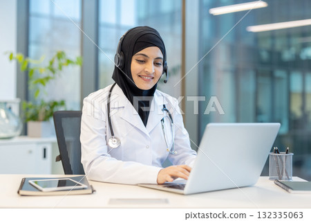 Smiling young muslim woman doctor wearing a headset and stethoscope, providing remote healthcare services and online medical advice using a laptop in a modern office 132335063