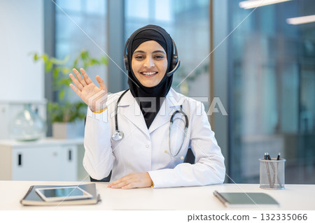 Muslim female doctor wearing medical uniform and stethoscope, smiling and waving hand during an online video call for a telemedicine consultation in a modern office Muslim female doctor wearing medical uniform and stethoscope, smiling and waving hand during an online video call for a telemedicine consultation in a modern office 132335066