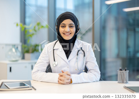 Confident muslim female doctor wearing headset smiling at camera during online consultation in bright modern medical office, providing telehealth services 132335070