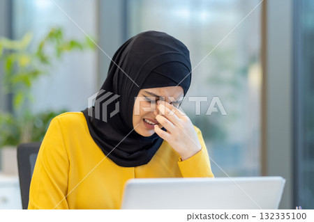 Young businesswoman wearing a hijab, rubbing her eyes in frustration while working on a laptop, dealing with headache and eye strain in a modern office environment Young businesswoman wearing a hijab, rubbing her eyes in frustration while working on a laptop, dealing with headache and eye strain in a modern office environment 132335100