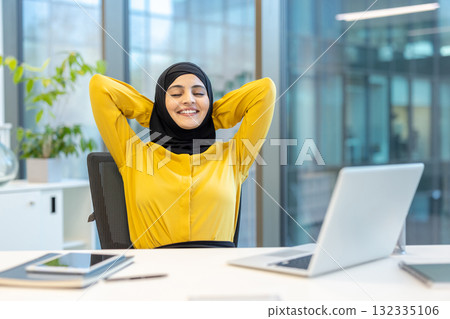 Muslim businesswoman is stretching and relaxing in her office chair, feeling great and happy with closed eyes and hands behind her head after a successful day of work Muslim businesswoman is stretching and relaxing in her office chair, feeling great and happy with closed eyes and hands behind her head after a successful day of work 132335106