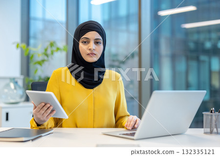 Young muslim businesswoman wearing a hijab and a yellow blouse, looking at the camera while working with a laptop and holding a digital tablet in a modern office environment Young muslim businesswoman wearing a hijab and a yellow blouse, looking at the camera while working with a laptop and holding a digital tablet in a modern office environment 132335121