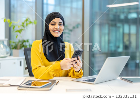 Muslim businesswoman smiling while using smartphone, sitting at office desk, reflecting confidence and professionalism in a contemporary workspace with a laptop and tablet ready for work 132335131
