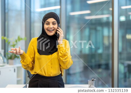 Muslim businesswoman wearing a hijab and yellow shirt smiling happily while having a phone conversation, making a hand gesture in a modern corporate office setting 132335139