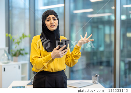 Muslim businesswoman wearing hijab, reacting with frustration and confusion while holding a smartphone and seeing bad news or a problem with modern technology in a corporate office setting 132335143