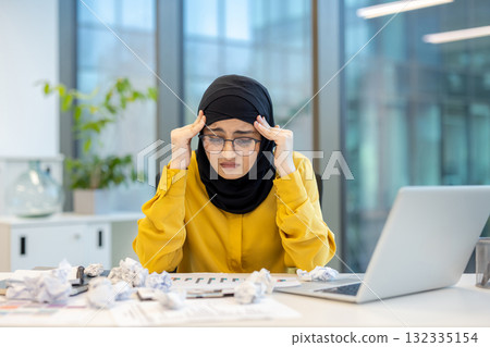 Young muslim businesswoman in hijab and glasses clutching her temples at a cluttered office desk, stressed and fatigued from overwork, missed deadlines and burnout 132335154