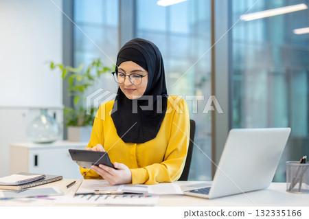 Muslim businesswoman wearing a black hijab and glasses, working in a modern office, concentrating while using a calculator for financial planning and accounting tasks on her desk Muslim businesswoman wearing a black hijab and glasses, working in a modern office, concentrating while using a calculator for financial planning and accounting tasks on her desk 132335166