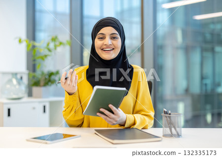 Muslim businesswoman in a black hijab and yellow blouse smiling confidently at her modern office desk while holding a notepad and pen, ready to plan and organize work 132335173