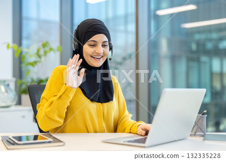 Professional muslim woman in hijab wearing a headset, smiling and waving during a video call on her laptop from a modern office workspace, confident and engaged in virtual customer service 132335228
