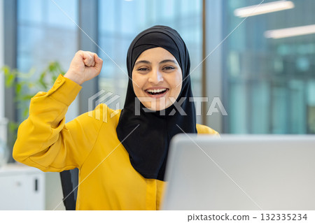 Muslim businesswoman wearing a hijab, cheering and showing a fist pump while working on a laptop in a modern office, celebrating achievement and triumph Muslim businesswoman wearing a hijab, cheering and showing a fist pump while working on a laptop in a modern office, celebrating achievement and triumph 132335234