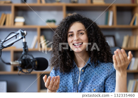 Happy young woman recording a podcast from home, speaking into a microphone and gesturing with her hands, surrounded by books on shelves in her cozy home studio Happy young woman recording a podcast from home, speaking into a microphone and gesturing with her hands, surrounded by books on shelves in her cozy home studio 132335460