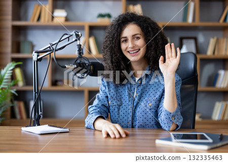 Woman with curly hair smiling and waving while recording a podcast or live stream from her home studio, engaging viewers with a confident, casual on-camera presence Woman with curly hair smiling and waving while recording a podcast or live stream from her home studio, engaging viewers with a confident, casual on-camera presence 132335463