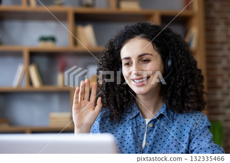 Woman in a headset smiling and waving at a laptop camera during a remote video conference, connecting with colleagues or friends from her home office setup Woman in a headset smiling and waving at a laptop camera during a remote video conference, connecting with colleagues or friends from her home office setup 132335466