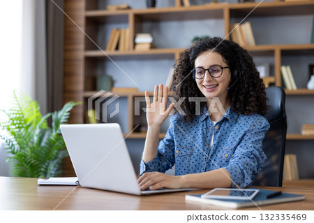 Young woman wearing glasses and headset smiling, waving hand while participating in a video call or online meeting on her laptop from a modern home office setting Young woman wearing glasses and headset smiling, waving hand while participating in a video call or online meeting on her laptop from a modern home office setting 132335469
