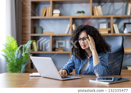 Young woman with curly hair and glasses feeling stressed and having a headache while using a laptop at home, representing challenges of remote work, pressure, and burnout Young woman with curly hair and glasses feeling stressed and having a headache while using a laptop at home, representing challenges of remote work, pressure, and burnout 132335477