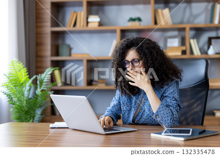 Stressed female entrepreneur covering her mouth with hand while reading shocking news on laptop, sitting at wooden desk in home office with bookshelves in background Stressed female entrepreneur covering her mouth with hand while reading shocking news on laptop, sitting at wooden desk in home office with bookshelves in background 132335478