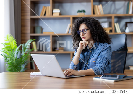 Young professional with curly hair and glasses concentrating on a laptop at a home office desk, focused on work or study with bookshelf and plant in the background Young professional with curly hair and glasses concentrating on a laptop at a home office desk, focused on work or study with bookshelf and plant in the background 132335481