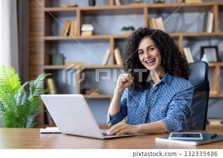 Smiling young woman with curly hair celebrates remote work success at her home office desk, using a laptop and looking at the camera with excited, confident joy and accomplishment Smiling young woman with curly hair celebrates remote work success at her home office desk, using a laptop and looking at the camera with excited, confident joy and accomplishment 132335486