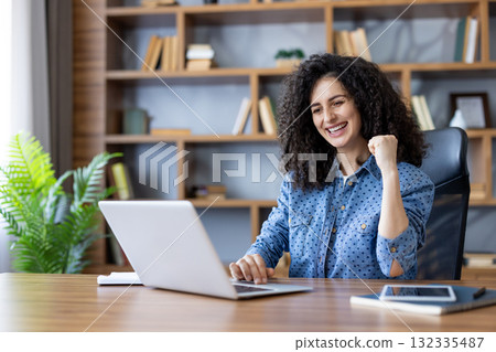 Happy curly-haired woman celebrates remote work success at a bright home office desk, smiling at her laptop with joy and a confident, productive energy Happy curly-haired woman celebrates remote work success at a bright home office desk, smiling at her laptop with joy and a confident, productive energy 132335487