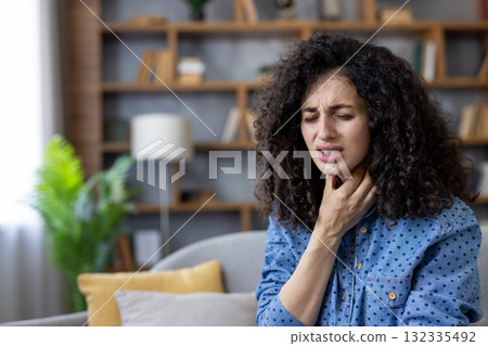 Young woman experiencing throat pain while touching her neck, sitting on a sofa at home, showing early signs of cold or flu with symptoms of tonsillitis or laryngitis Young woman experiencing throat pain while touching her neck, sitting on a sofa at home, showing early signs of cold or flu with symptoms of tonsillitis or laryngitis 132335492