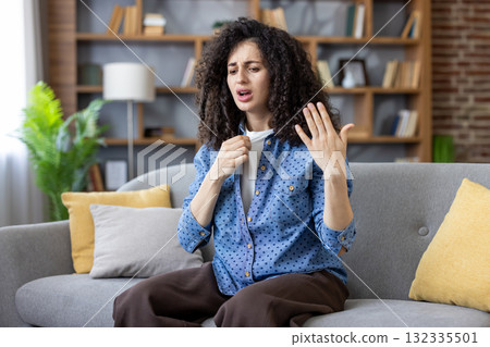 Young woman sitting on sofa at home, trying to cool herself down, suffering from high temperature and hot weather, feeling unwell, experiencing discomfort from summer heatstroke 132335501