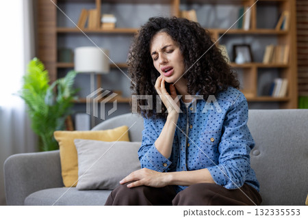 Young woman sitting on the sofa at home, experiencing intense toothache, pressing her hand against her cheek and grimacing in response to the overwhelming pain 132335553