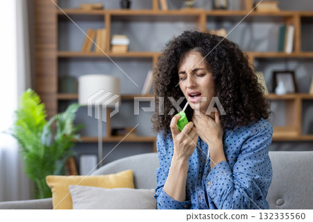 Young woman on a couch spraying throat medicine into her mouth, wincing in discomfort as she self-treats sore throat and cold symptoms at home Young woman on a couch spraying throat medicine into her mouth, wincing in discomfort as she self-treats sore throat and cold symptoms at home 132335560