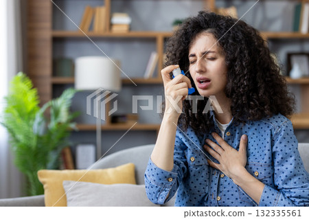 Young woman sitting on a sofa at home, using an inhaler during an asthma attack, struggling to breathe, holding her chest in discomfort and facing a respiratory crisis Young woman sitting on a sofa at home, using an inhaler during an asthma attack, struggling to breathe, holding her chest in discomfort and facing a respiratory crisis 132335561