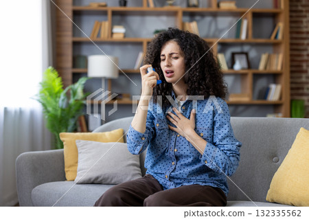 Young woman having difficulty breathing and managing an acute asthma attack at home, using an inhaler to alleviate respiratory symptoms and seeking quick relief Young woman having difficulty breathing and managing an acute asthma attack at home, using an inhaler to alleviate respiratory symptoms and seeking quick relief 132335562