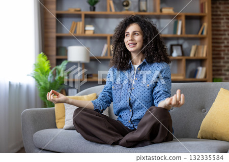 Woman with curly hair sitting in a lotus position on a living room sofa, practicing meditation and mindfulness for mental health and relaxation at home, enhancing her overall wellbeing Woman with curly hair sitting in a lotus position on a living room sofa, practicing meditation and mindfulness for mental health and relaxation at home, enhancing her overall wellbeing 132335584
