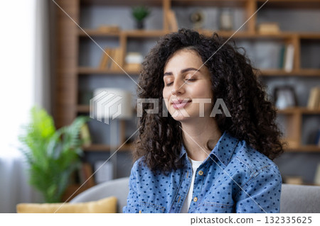 Woman with curly hair closing eyes meditating, practicing mindfulness, feeling peaceful and relaxed while sitting in the comfort of her living room, focusing on self-care and well-being 132335625