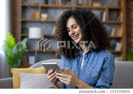 Woman with curly hair smiling and looking at a tablet, sitting on a sofa at home, enjoying online content and connectivity for entertainment and relaxation 132335643
