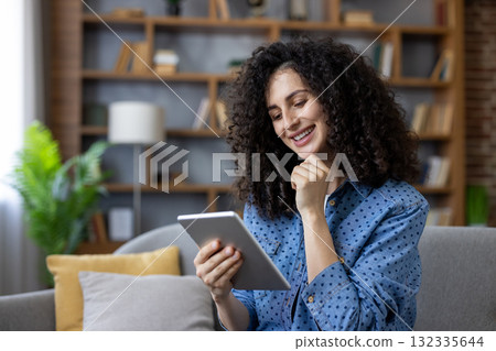 Happy young woman with curly hair relaxing on a sofa, smiling while browsing content on a digital tablet, enjoying leisure time in a modern living room with a bookshelf Happy young woman with curly hair relaxing on a sofa, smiling while browsing content on a digital tablet, enjoying leisure time in a modern living room with a bookshelf 132335644