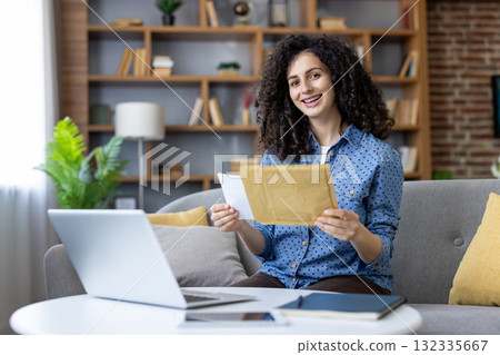 Young woman with curly hair smiling as she opens an envelope on a sofa beside her laptop in a cozy home office, receiving happy personal or work-related mail Young woman with curly hair smiling as she opens an envelope on a sofa beside her laptop in a cozy home office, receiving happy personal or work-related mail 132335667