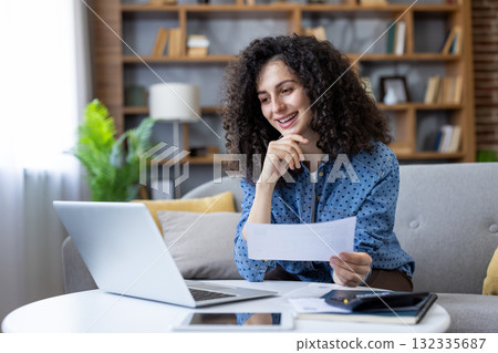 Smiling female freelancer reading good news in a letter while working remotely on a laptop, managing domestic finances and enjoying flexible work arrangements 132335687